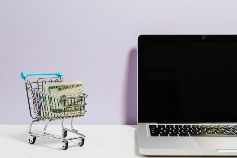 A tiny shopping cart with some cash in it next to a laptop, representing customer retention strategies to try in 2021